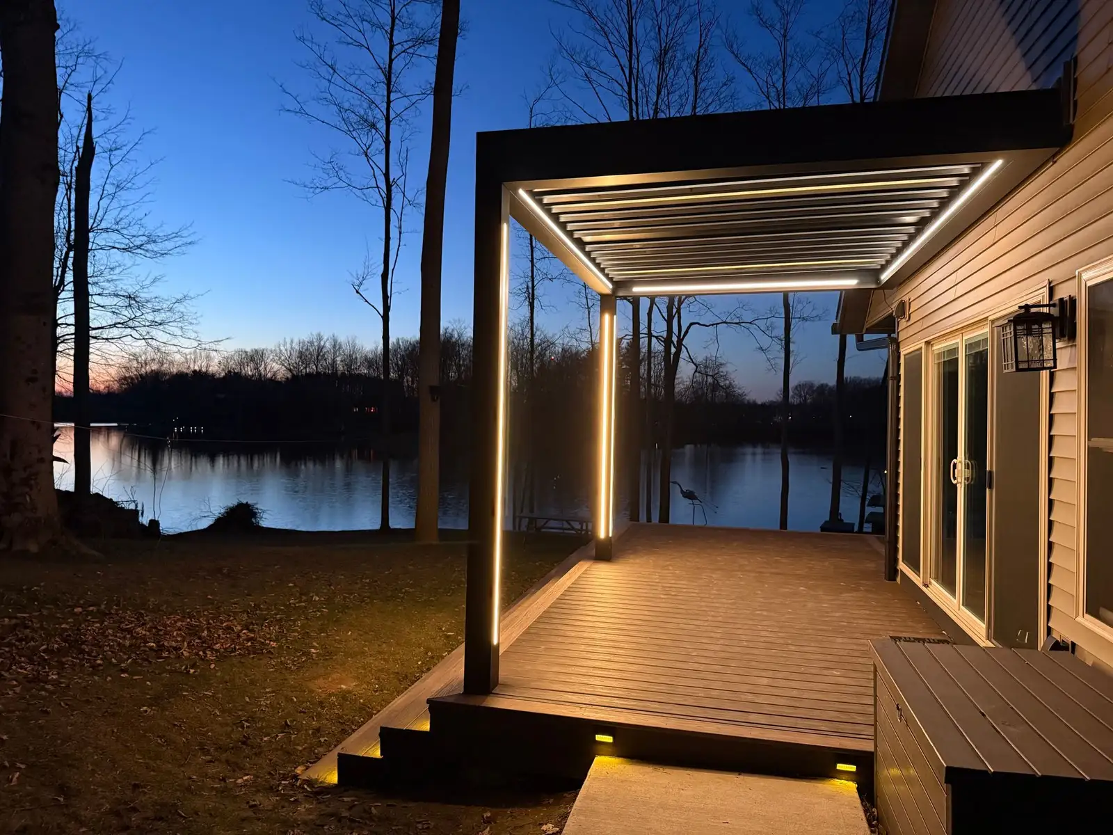 Black louvered pergola with warm LED strip lighting over a composite deck, looking out to a lake at dusk