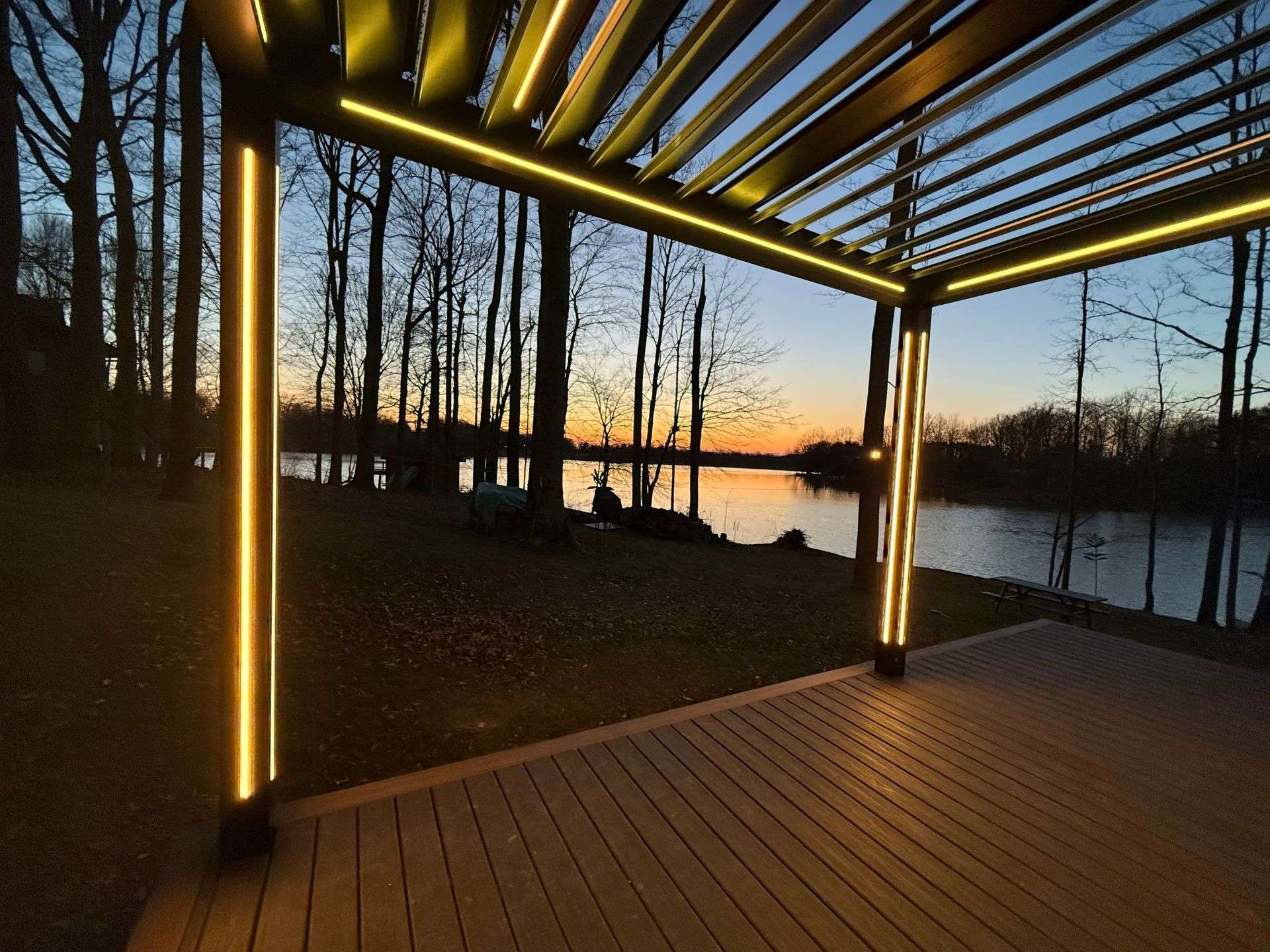 View from under a lit louvered pergola across a deck to a lake at sunset